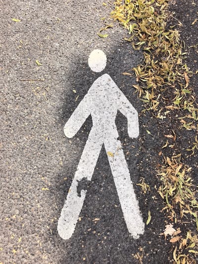 The image shows a white pedestrian crossing symbol painted on dark asphalt. The symbol depicts a stylized figure with a round head, a rectangular torso, and two angled legs. The figure is oriented vertically, facing upwards towards the top of the frame. The painting is somewhat worn, with some of the white paint chipped and revealing the black asphalt beneath. Small bits of dried leaves, some green and some yellow, are scattered around the symbol and on the asphalt, particularly to the right of the figure. A thin twig is also visible on the asphalt to the left of the figure's legs. The scene is an outdoor setting, likely a pathway or road in Janvry, France. The lighting suggests it is daytime, possibly sunny, as there is a slight shadow cast on the asphalt to the right of the figure. There is no visible text in the image.