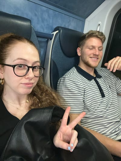 This image depicts two individuals seated within the interior of a public transportation vehicle, consistent with a train, presumed to be operating in or around Newark, United States. In the foreground on the left, a female with long, curly reddish-brown hair and black-rimmed glasses looks directly at the camera, her left hand displaying a peace sign (V-gesture) with light blue painted fingernails, resting on a black leather-like bag. She wears a black top, a gold hoop earring on her left ear, and layered gold necklaces, one with a small heart pendant. To her right, a male with short light brown hair and light facial hair smiles with closed eyes. He wears a white polo shirt with horizontal dark stripes and a dark collar. A gold-colored ring is visible on the fourth finger of his right hand. The vehicle's interior features dark blue fabric seats with grey plastic frames and blue upper wall paneling. A dark window is visible on the right side of the frame.