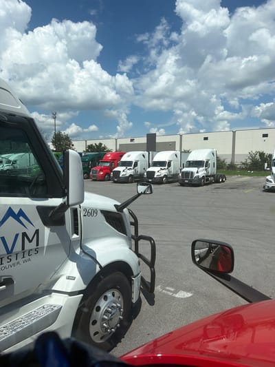 A fleet of white and red semi-trucks are parked in a lot with a warehouse in the background under a partly cloudy sky. The trucks are arranged in rows, with some on the left side of the image in the foreground and middle ground, and others in the middle and right ground. The truck in the immediate foreground on the left is white with blue and black lettering that says "VM LOGISTICS VANCOUVER, WA". A red semi-truck is parked to the right of the foreground truck, and further back, several more white and red semi-trucks are visible. The warehouse in the background is a large, beige structure with dark trim and windows. The ground is paved asphalt, and there are some trees and bushes visible in the distance. A traffic light is visible in the background.