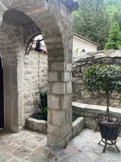 The image shows a stone archway with a bell hanging from its interior. The archway appears to be part of a building or structure made of stone blocks, suggesting historical construction. A potted plant is positioned on a stone surface nearby. The archway is located in Podličak, Montenegro.