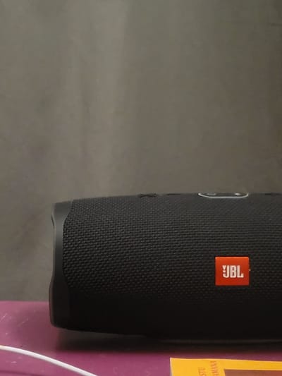 The image shows a black JBL portable speaker resting on a purple suitcase.  The image is taken from a low angle, looking up at the speaker.  The speaker is in focus, while the background is out of focus. The speaker is made of a black fabric material with a white JBL logo on the side.  The image was taken in Calais, France at the coordinates 50.948, 1.846.