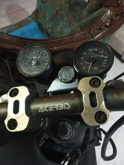A close-up, eye-level photograph depicts the dashboard and handlebars of a motorcycle, positioned indoors in the city of B. Nam Ngao, Laos. The central focus is the instrument cluster, featuring two circular gauges and an ignition switch.
The left gauge, a tachometer, displays "MR. D" centrally and "x1000rpm" at the bottom, with a scale ranging from 0 to 12. Its needle rests at zero. The right gauge, a speedometer, shows a scale from 0 to 160 km/h, with an odometer reading of 17098 km visible. Its needle is also at zero. Both gauges appear aged and show signs of dust or discoloration on their faces.
Between the two gauges is a circular ignition switch, exhibiting wear around the keyhole. Below the instrument cluster, a dark-colored handlebar is visible, bearing the white "ACERBIS" logo and subtle numerical markings. The handlebar is secured by two prominent, gold-toned or brass-colored clamps with visible bolts.
The background is out of focus, revealing what appears to be a portion of ornate wooden furniture with carved details, possibly a chair, upholstered in a pattern of teal and brown/gold floral designs. A segment of bright blue fabric is visible draped over the furniture. No individuals are present in the scene.