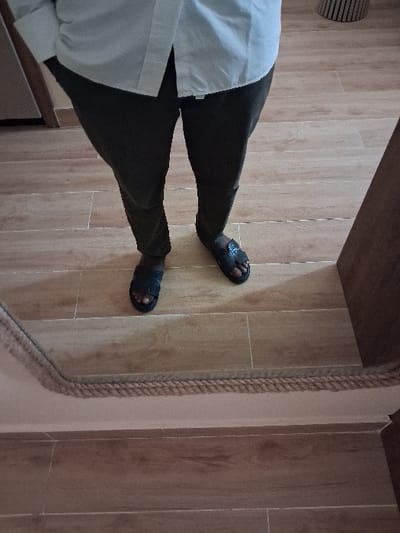The image, captured in the city of Takum, Nigeria, displays a person from the waist down, reflected in a mirror. The individual wears a white long-sleeved shirt and dark-colored trousers, with hands in the pockets. On their feet are black open-toed sandals. They stand on a light-toned ceramic tiled floor, which features a wood-grain pattern laid in a staggered bond. The lower edge of the mirror is visible in the foreground, framed by a braided rope-like border. Reflected in the background are a partial wooden door frame to the left and a vertically striped object, consistent with a basket or stool, to the right.