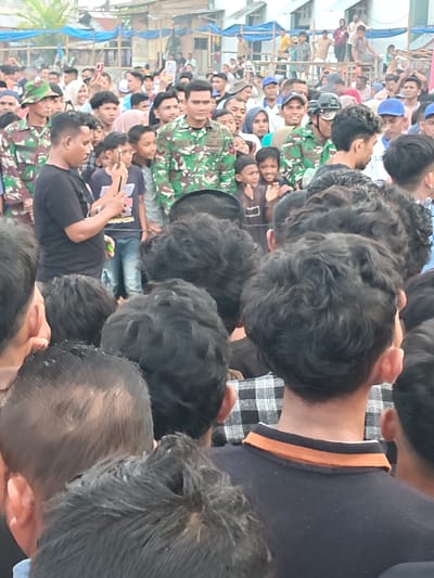 The image captures a lively outdoor gathering in Desa Meunasah Blang, Indonesia. A crowd of people, predominantly young men and boys, are densely packed together, suggesting a significant event or popular attraction. Many are looking in the same direction, with some holding up smartphones, possibly to capture the moment or a performance.
In the center, a man in a camouflage uniform, likely a soldier, stands out. He is the focal point for some of the onlookers. The atmosphere appears energetic and engaged. The time of day seems to be daytime, with natural light illuminating the scene. The background reveals a mix of structures, including what appear to be temporary shelters or stalls, perhaps indicating a local market, festival, or community event. The presence of blue tarps suggests some form of temporary roofing or shading.
The clothing of the individuals is varied, from casual t-shirts to the military uniform. The hairstyles are diverse, with many young men sporting curly or short cuts. The overall impression is one of a busy public assembly, possibly a cultural celebration, a gathering of community members, or a military-related event drawing local attention. No specific text is clearly legible in the image.