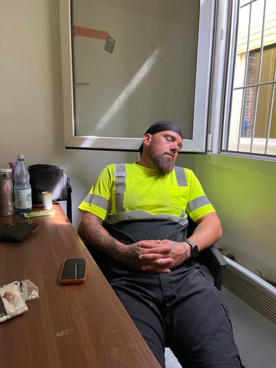 A man in a neon yellow and gray high-visibility shirt, dark pants, and a black skullcap is asleep, seated in a black office chair next to a window. His head is tilted down, eyes closed, and hands are clasped in his lap. The window has a metal grate on the outside. On a wooden desk to the man's left are a bottle of water, a small can, a black bag, and a smartphone. The scene appears to be an office or break room, possibly in Neuss, Germany.