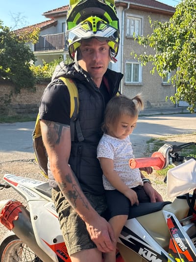 In Oreshak, Bulgaria, a man wearing a yellow and black motocross helmet and a black puffer vest over a black t-shirt is seated on a white and orange dirt bike. He has a yellow backpack on. A young child with a ponytail is sitting in front of him on the dirt bike, wearing a white t-shirt with a repeating pattern of small, light-colored objects. The child is looking down. The man has tattoos on his left arm, including a roman numeral and an arrow design. The dirt bike has "LIFEPROOF" and "KTM" branding. The background shows a house with a balcony and trees, illuminated by sunlight.