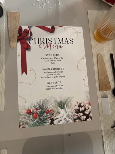 This close-up image captures a festive table setting, likely prepared for a Christmas meal in Bellevue, Switzerland. The central focus is an elegantly designed "CHRISTMAS Menu" card, positioned diagonally on a light grey table surface. The menu features holiday-themed graphics including a red ribbon and bow, golden stars, and frosted pine branches with red berries and pinecones, creating a winter holiday atmosphere.
To the upper left of the menu, a ribbed glass holds an amber-colored liquid. Above the menu, a light wooden spatula or serving utensil is visible, alongside a small white rectangular object and part of a black utensil handle, all resting on a light-colored woven placemat. On the lower left, shiny, flaky decorative elements, resembling artificial snow or glitter, are scattered on the table.
The visible text on the menu, presented in French, details the meal courses:
* **STARTERS:** "Chips nature & paprika" (plain and paprika crisps), "olive verte / maïs" (green olives / corn), and "flute" (likely a type of bread or cracker).
* **MAIN COURSES:** "Raclette au fromage" (cheese raclette), "viande séchée" (dried meat), and "cornichon et petit oignon" (gherkins and small onions), clearly indicating a traditional raclette dinner.
* **DESSERTS:** "Gâteau au chocolat" (chocolate cake) and "Sapin en chocolat" (chocolate Christmas tree).
The scene depicts a thoughtfully arranged setting for a holiday feast, evoking a warm and celebratory mood, characteristic of Christmas gatherings.