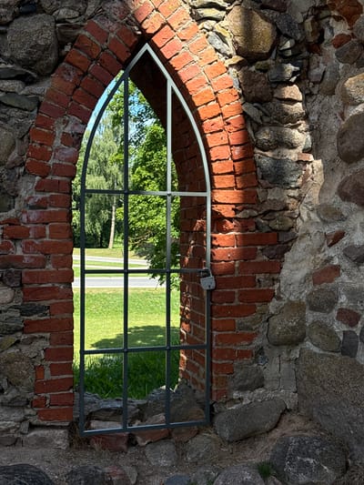 An arched brick opening in a stone wall features a metal gate with a padlock. Through the opening, a grassy area with trees and a road is visible. The scene is illuminated by sunlight casting shadows.