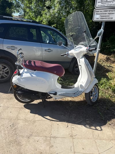 This media file shows a white scooter parked next to a light-colored car on a sunny day in Dyuni, Bulgaria. The scooter is parked in front of a pole with signs. One sign is in Bulgarian and appears to be related to parking enforcement, listing phone numbers and indicating "Nakazatelen parking" (Penalty parking) and "Primorsko" (a nearby town). The scooter has a large windscreen and a dark red seat. The car is partially visible, with its side door and windows showing. The ground is a mixture of dry grass and paved surfaces, with tire tracks and shoe prints visible on the pavement. The sun casts a long shadow of the scooter onto the ground, indicating it is likely late afternoon or early morning. The overall scene suggests an outdoor parking area, possibly near a tourist destination given the parking information.