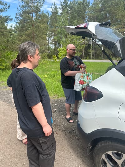 The image shows two men standing near a white car with its trunk open. One man, bald and wearing glasses, is taking a patterned bag out of the trunk. He is wearing a black t-shirt and shorts with sandals. The other man, with graying hair and a ponytail, is standing nearby, facing the other man. He is wearing a black t-shirt and dark-colored pants. The car is parked on a paved surface, possibly a road or parking area. Trees and greenery are visible in the background, suggesting a rural or suburban setting. The location is Юзуфоўскі сельскі Савет, Belarus.