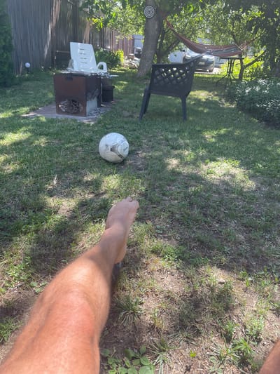 The image depicts a backyard scene on a sunny day. In the foreground, a person's bare leg and foot are visible, suggesting they are sitting or lying on the grass. A white soccer ball with black accents rests on the lawn a short distance away. In the middle ground, a dark plastic chair is positioned on the grass, and behind it, a hammock is strung between trees. A small metal table with a black top is next to the hammock. Further back, a barbecue grill with a white plastic chair on top is visible near a wooden fence. The environment is a yard with green grass, trees, and dappled sunlight. The image does not contain enough information to determine the specific location in Krutets, Russia.