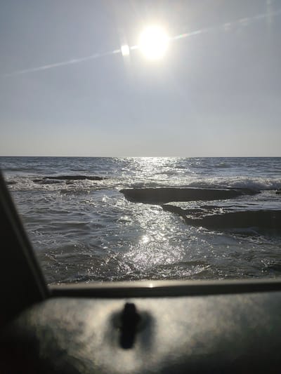 This media file is a photograph of a seascape with the sun shining brightly in the sky. The scene depicts a body of water, likely the sea, with waves gently rolling onto a rocky shore. The sun, positioned high in the sky, creates a brilliant glare and reflects off the water's surface, casting shimmering highlights. The sky is clear and pale blue, with a thin, elongated cloud streak visible to the left of the sun. In the foreground, out of focus, is a dark, metallic object with a visible bolt or screw, suggesting the photograph may have been taken from a boat, a pier, or some other structure overlooking the water. The lighting suggests it is daytime, likely late morning or early afternoon, given the sun's high position and the intensity of its rays. The overall mood is serene and peaceful, with the natural beauty of the ocean and the bright sunlight creating a calming atmosphere. There is no visible text in the image.