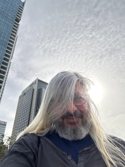 A low-angle outdoor shot depicts an individual against a backdrop of modern high-rise buildings under an overcast sky. The subject, a male, fills the lower half of the frame, featuring long, light grey hair that partially obscures his face, a full beard of similar coloration, and round-framed glasses. He wears a dark, possibly charcoal-grey, collared shirt over a navy blue undershirt, and is smiling faintly. The sun, positioned behind and to the right of the subject's head, appears as a bright glare.
In the background, to the left, a tall, glass-fronted skyscraper with numerous balconies is visible, with the Hebrew text "אֶלעל" (El Al) partially visible near its base. To the right, another prominent dark-glass skyscraper is clearly labeled "BSR 2" near its top. The sky exhibits a textured pattern of altocumulus clouds. The location is identified as the Bnei Brak Business Center, Bnei Brak, Israel, based on the visible BSR 2 tower.