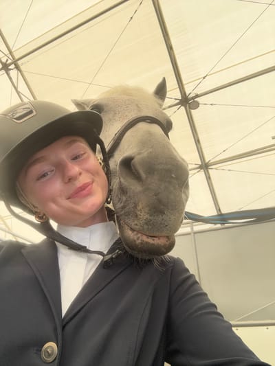 A forensic close-up image depicts a young female and a light grey horse within an indoor equestrian facility, consistent with locations found in the Township of Langley, Canada. The female, positioned on the left, wears a dark equestrian helmet, a white collared shirt, and a dark blue riding jacket with a circular gold button. She displays a subtle smile and looks directly forward. The horse, equipped with a dark leather bridle, has its muzzle and lower jaw resting on the female's right shoulder. Its eyes are partially visible, looking slightly left. The background consists of a light-colored fabric ceiling, exhibiting a structural framework of metal beams and taut cables, characteristic of a large tent or covered arena. Diffused light illuminates the scene from above.