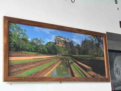 This is a horizontal, framed photograph of a historical site featuring a large rock formation in the background and meticulously maintained gardens and water features in the foreground. The rock formation, identified as Sigiriya or Lion Rock, is a prominent natural landmark with a distinct flat top, characteristic of the ancient royal palace and fortress complex located in the Matale District of Sri Lanka, near the town of Dambulla. The foreground displays a series of terraced gardens and brick-lined water channels, some of which are filled with calm, reflective water. Lush green grass covers parts of the terraces, contrasting with the weathered red bricks. A group of people, small in scale relative to the landscape, are walking along a pathway in the middle ground, suggesting the site is accessible to visitors. The sky is a clear blue with scattered white clouds, indicating a bright day. The photograph is framed with a wooden border. The image is likely a souvenir or decorative piece, intended to showcase the natural and historical beauty of the location.