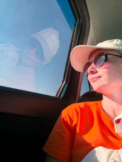 The image shows a young woman wearing sunglasses and a white cap, looking out of the window of a vehicle. She is wearing an orange t-shirt. Her reflection is visible in the window. The scene appears to be outdoors on a sunny day. The location is specified as Karaiganovo, Russia.