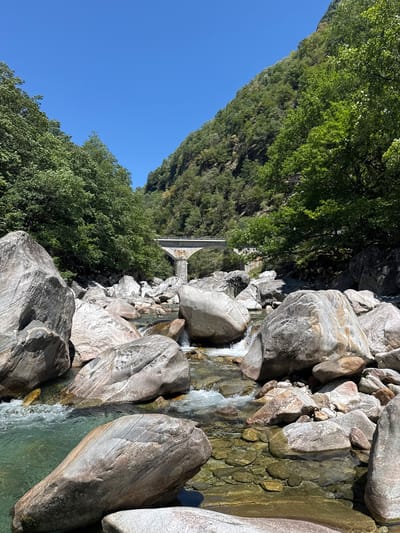 The image depicts a clear, shallow river flowing through a rocky gorge. The riverbed is composed of smooth, rounded stones of various sizes, with some areas of sand and gravel. The water is a vibrant turquoise color, and its clarity allows for a clear view of the submerged rocks. Large, grey boulders are scattered along the riverbanks and within the water, creating an uneven and rugged landscape. Lush green trees and vegetation cover the steep slopes of the gorge, contrasting with the grey rock and blue sky. In the distance, a multi-arched stone bridge spans the gorge, suggesting a man-made element within the natural scenery. The sky is a bright, cloudless blue, indicating a sunny day. The overall scene is one of natural beauty and rugged terrain. This is likely the Verzasca River in Ticino, Switzerland, famous for its clear turquoise waters and surrounding natural landscape.