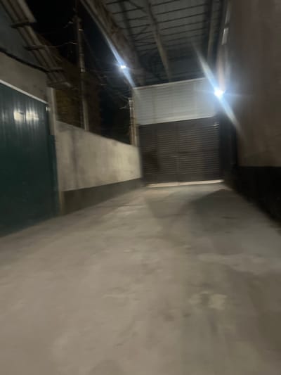 The image depicts an outdoor scene, possibly at night, with a concrete ground and walls on either side. A large, dark-colored metal gate is visible at the end of a pathway, with a white structure above it. To the left, a dark green metal fence or door runs along a concrete wall. The overall lighting is dim, with bright light sources causing lens flare and obscuring details. The scene appears to be a pathway or alleyway. There are no people or specific actions visible in the image. The location is stated to be in Lebedinovka, Kyrgyzstan.
