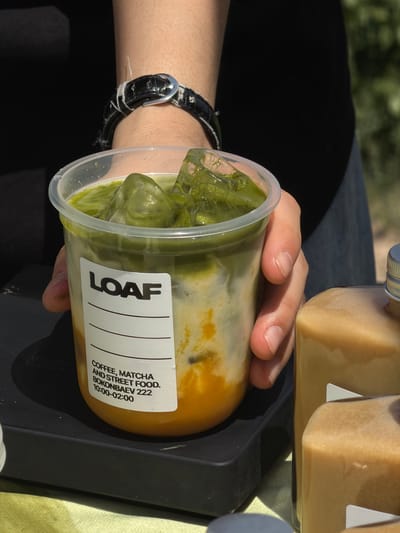 A person is holding a transparent plastic cup containing a beverage with green ice cubes. The cup has a white label with the word "LOAF" in bold black letters, followed by "COFFEE, MATCHA AND STREET FOOD. BOKONBAEV 222 10:00-02:00" printed below. The beverage appears to have multiple layers of different colors. The person is wearing a black watch with a silver buckle on their wrist. A black tray supports the cup and a bottle with a light brown liquid. The scene is outdoors, with the sun illuminating the objects, suggesting daytime. The location, according to the cup's label, is Bokonbaev 222, indicative of Kok Jar, Kyrgyzstan.