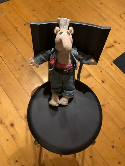A stuffed toy with tan fur, large black eyes, a long tan snout, and grey tufts on its head stands on a black chair. The toy is wearing a denim jacket over a pink belt and blue denim pants. A red circular pin with a white "W" is attached to the left side of the jacket. The chair is positioned on a wooden floor. The location is Mepiskalaki, Georgia.