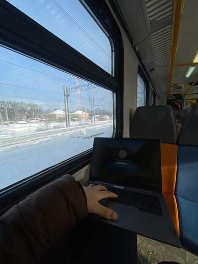 An interior view from a train carriage depicts a snowy landscape outside the window, located near Fryazevo, Russia. A person, whose right arm in a brown jacket sleeve and hand with a ring are visible, operates a dark grey laptop resting on their lap. The laptop screen displays a login prompt featuring a user profile icon and the name "Victoria Zhulyabina." Outside the window, a clear blue sky spans over snow-covered ground and railroad tracks. In the midground, several light-colored buildings are visible among snow-dusted trees, with a distinctive red and white lattice telecommunications tower in the distance. Overhead power lines for the electrified railway cross the frame. Inside the train, light-colored walls, ribbed ceiling panels, and portions of grey and orange/blue upholstered seats are discernible, along with yellow grab handles and overhead lighting.