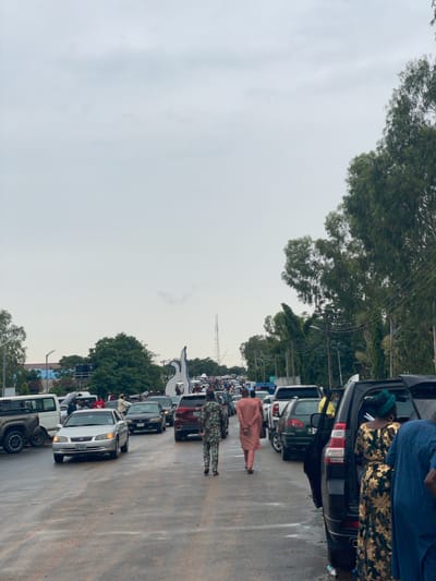 The image depicts a traffic jam on a road in New Nyanya, Nigeria. Several vehicles are lined up, suggesting a considerable delay. People are visible standing or walking near the vehicles, some dressed in traditional attire, including a person in a peach-colored outfit and another in a patterned green and white garment. A black SUV with its trunk open is prominent in the foreground, with individuals standing beside it. The sky is overcast, indicating cloudy weather, and the lighting suggests it is daytime, possibly late afternoon or early evening. Trees line the side of the road, and power lines are visible overhead, typical of an urban or semi-urban environment. The scene appears to be one of inconvenience and waiting, with no explicit signs of distress or urgency, suggesting a common occurrence of traffic congestion in the area.