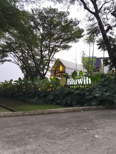 This image captures an exterior view of "The Kluwih Heritage" in Babakan Madang, Indonesia, likely during the late afternoon or early evening. No people are visible in the frame.
In the foreground, a dark, textured asphalt or concrete path leads up to a low concrete barrier. Beyond the barrier, a strip of green grass gives way to dense, lush green foliage. A prominent, illuminated sign is partially nestled within these plants, clearly displaying the text "THE KLUWIH HERITAGE" in bright white letters. Scattered warm lights are visible among the plants, casting a soft glow.
In the mid-ground and background, modern architectural structures with white corrugated roofs and large window sections are visible, resembling contemporary barn-style buildings. One building, positioned centrally, has large glass panels that reflect a warm interior light. These structures are surrounded and partially obscured by numerous tall, mature trees with dense green canopies, some of which appear to be palm trees. The trees dominate the upper portion of the image, silhouetted against a bright, overcast or twilight sky, suggesting a serene and natural setting for the establishment. The scene conveys a tranquil and welcoming ambiance.