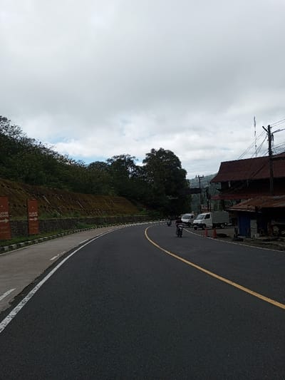 The image displays a curving asphalt road in Phoxay, Laos, under an overcast sky. The road features a solid yellow center line and a white line marking the right shoulder, alongside a paved pedestrian path. To the left of the road, a concrete retaining wall, partially covered in moss, supports a steep hillside densely covered with green trees and vegetation. Attached to this wall are multiple orange banners, some visible reading "SLOW DOWN" and "LAKE VIEW RESORT" with directional arrows.

On the road, two individuals are visible riding motorcycles, traveling away from the viewer. Further down the road on the right, a white van and a light-colored utility truck are parked. Adjacent to these vehicles stands a two-story building with a prominent red-brown corrugated metal roof. Smaller, temporary-looking structures with blue and brown coverings are positioned in front of this building. Utility poles with numerous overhead wires are present along the right side of the road. An orange traffic cone is visible on the ground near the parked vehicles. In the background, beyond the buildings and vehicles on the right, a faint bridge structure or elevated roadway is discernible amidst distant hills.