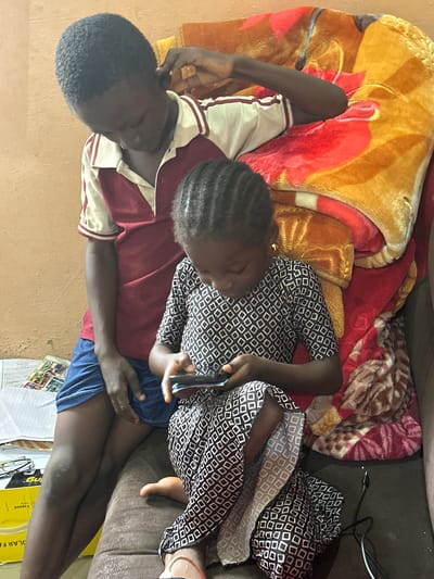 In Gidan Waziri, Nigeria, two Black children are seated on a couch covered with a vibrantly patterned blanket. The older child, positioned on the left, wears a maroon and white collared shirt and blue shorts. This child is leaning back, looking down, with their right hand touching their ear. The younger child, on the right, is engrossed in a smartphone held in their hands, wearing a black and white patterned dress. The background features a plain wall. Adjacent to the couch, a stack of papers and a yellow box are visible on the floor. A black cable is also present on the floor.