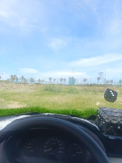 The image, likely taken from inside a vehicle, presents a view of a park or green space in Martil, Morocco. A strip of greenery with trees runs horizontally across the center of the frame. In the foreground, parts of the car's interior are visible, including the dashboard and steering wheel. Outside, the sky is light blue with scattered clouds. A fuzzy dice hangs from the rearview mirror.