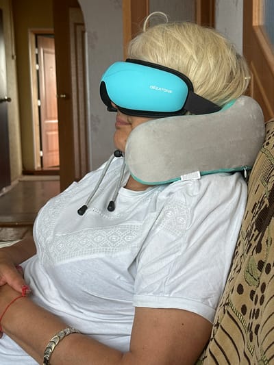 In Urazovo, Russia, a woman with blonde hair is seated on a sofa, wearing a white shirt and a blue eye mask. A grey neck pillow is around her neck, supporting her head and the eye mask. The eye mask has the brand name "Gezatone" printed on it in white lettering. She has a bracelet on her left wrist and a red string tied around her right wrist. The background shows a doorway and a wall with wallpaper. The lighting suggests it is daytime.