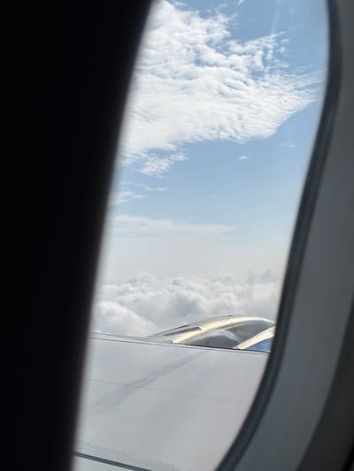 The image is a view from an airplane window, showing clouds and the wing of the aircraft. A dark, solid object, likely part of the airplane's interior, occupies the left side of the frame, obscuring a portion of the view. The sky is blue with scattered white clouds. The wing of the airplane is white and metallic, with visible rivets along its edge. A portion of what appears to be an engine cowling, with a shiny, metallic finish, is visible above the wing. The perspective suggests the photograph was taken while in flight. There is no indication of Mohan, India, or any urban environment in the image.