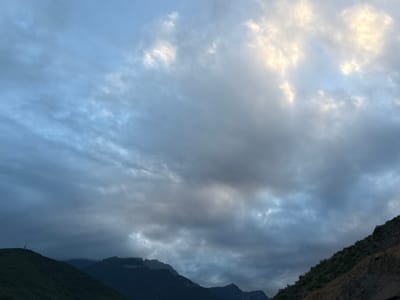 The image captures a dramatic sky over a mountainous landscape. The sky is filled with a mix of dark, heavy clouds and patches of bright, luminous clouds, suggesting a dynamic weather system or the approach of dawn or dusk. The lighting is diffused, with some areas strongly illuminated by what appears to be the sun filtering through the clouds, creating a soft glow.

In the foreground, the dark silhouettes of mountains dominate the lower portion of the frame. The slopes of the mountains are rugged and appear to be covered in vegetation, though the details are obscured by the low light. On the left, a steep, dark green mountain slope rises, topped by a thin, dark structure resembling a tower or antenna. The right side of the image features a steep, rocky incline with sparse vegetation. The overall impression is one of a remote, natural setting.

The time of day is likely early morning or late evening, given the low light and the quality of the illumination from the clouds. The weather appears to be overcast or transitioning, with a sense of atmosphere. The location, Kapan, Armenia, suggests a mountainous region. There are no visible people or text in the image. The mood conveyed is one of awe and perhaps a little somberness, due to the dark clouds and stark landscape.