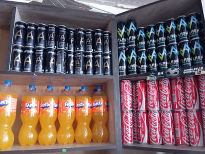 In an indoor setting, possibly a store or pantry, a variety of beverages are displayed on shelves. The left side of the image features rows of Coca-Cola cans, some with "FIFA WORLD CUP" branding and "CLASSIC" labels. To the right of the Coca-Cola cans are rows of energy drinks, including "FLASH" energy drinks with a distinctive lightning bolt logo and "Adrenaline Rush" energy drinks with a prominent "A" logo. The far right of the image showcases several large bottles of Fanta, an orange-flavored soda, with clear blue caps. The lighting appears to be artificial, suggesting an indoor environment. No people are visible in the frame, and the scene depicts a well-stocked display of popular drink brands. Visible text includes brand names, product descriptions like "CLASSIC," "FIFA WORLD CUP," "ENERGY," and "RUSH," and volume indicators like "1.5 L." The overall impression is a retail or storage area focused on beverage sales.