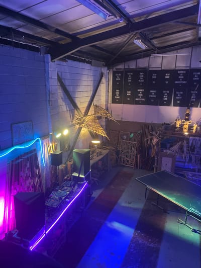 The image shows an interior space with exposed brick walls, a high ceiling with industrial metal beams, and various objects and installations. There's a large, intricately crafted sculpture, possibly made of reeds or straw, lit by bright lights. Signs with text are hung on the wall. A ping pong table is present and the scene is illuminated with neon lights, creating a mix of industrial and artistic elements. The overall atmosphere appears to be experimental and creative.