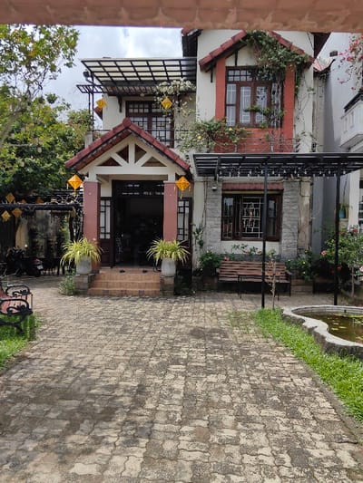A two-story building, located in Phú Xuân Ward, Vietnam, is viewed from a paved courtyard under an overcast sky. The structure features white-painted walls with reddish-brown tiled gables and roof elements. The central entrance is accessed by three reddish-brown steps, flanked by two gray planters containing vibrant light-green foliage. The doorway is dark and open. Two square, yellow lanterns hang from the triangular-shaped tiled eaves above the entrance.

To the immediate right of the entrance, the ground floor section exhibits a stone-patterned facade with a dark-framed window. A plain wooden bench is positioned against the wall beneath a black metal pergola, from which multiple small white lights are suspended. A security camera is mounted on the wall above this window. The upper floor displays multiple dark-framed windows, with significant green and white-flowering plant growth cascading from a small balcony railing and around the window frames.

The foreground is dominated by a path paved with light-colored, irregularly shaped interlocking concrete pavers, showing areas of discoloration and wear. A narrow strip of green grass borders the paved path on the right, alongside an irregularly shaped, stone-edged pond containing murky water and some surface vegetation. To the far left of the paved area, a dark wooden bench with black metal scrollwork arms is partially visible on the grass. Further left, behind a weathered wall, two dark motorcycles are parked amidst dense green foliage. Additional square yellow lanterns and small white lights are strung among the trees and overhead structures on the left side of the scene. No individuals are present in the frame.