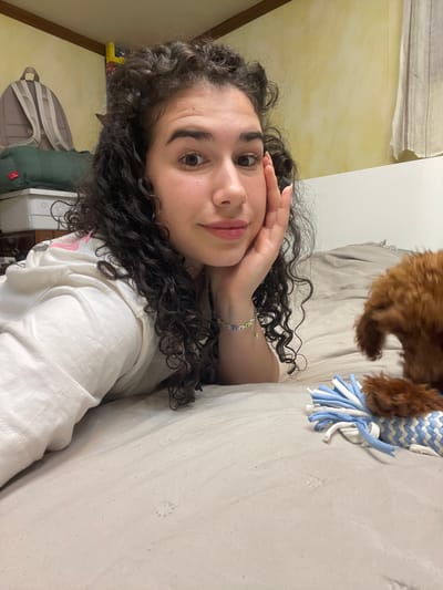 A candid, tilted portrait shows a young woman with dark, curly hair resting on her side, looking directly at the camera with a soft smile. She wears a light-colored hooded top and a beaded bracelet on her left wrist, adorned with green, blue, and white beads. Her right hand is gently placed near her forehead.
In the upper right portion of the frame, a small, fluffy brown dog is partially visible, appearing to interact with a blue and white patterned dog toy with frayed ends, which rests on a light-colored, textured surface, likely a bed or couch.
The indoor setting suggests a domestic environment, possibly a bedroom in Челопечене, Bulgaria. The background features a light yellow-cream textured wall with a wooden trim on the left, and a plain white wall or headboard on the right. Below the woman, a light-colored backpack, a dark green puffy bag, and a white electronic device are visible, further indicating a personal space. The lighting is soft and natural, suggesting a relaxed daytime atmosphere. No visible text is present.