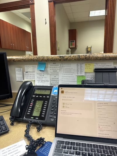 The image captures a detailed view of a desk area, likely in a medical or administrative office, looking across the countertop into a room beyond. The perspective is from above and to the right, showing various work-related items.

In the background, an interior room is visible through an archway framed with dark wooden trim. The room features light-colored walls and a drop ceiling with rectangular fluorescent light panels. A brown wooden cabinet with a lighter top and a colorful item (possibly a picture or sign) on it stands in the distance. To its right, some metal equipment or shelving is faintly discernible. Another long, low wooden cabinet is situated closer to the foreground, beneath the archway.

The desk surface in the foreground is a speckled, granite-like material with shades of brown, beige, and gray. Adhered to the vertical surface behind the desk, which appears to be a wall or partition, are numerous white papers and colorful sticky notes. These documents contain various pieces of handwritten and printed information. Visible text on the sticky notes includes:
*   Yellow notes: "TSPOT - 37737", "Hep B Antigen (Labcorp)", "032104", "866 95", "5054", "BOBS PAS".
*   Blue notes: "Cign", "543", "Kiemes", "800", "882", "446".
The white papers list several company names, account numbers, and contact details, suggesting affiliations with medical or lab services:
*   "QUEST Account #10081893", "ADD lab #866 697 8371", "Rep: Alexander Jimene".
*   "LABCORP Account 4212922".
*   "Alliance MRI Pearl P: 832-344-0008 F:832-344-0009".
*   "Houston Neurological Instite Phone: 281 922 7222 10905 Mononal Hemm TX 77584 For 713 947 6102".
*   "CPL Account 91007", "ADD 888-494-127", "Rep. Catia King".
*   "Braso Sport hospi 7285 114".
Some papers also show checklists with options such as "Selection of drug block list", "drug screen", "verify responsibility code", and "request approval from manager".

On the desk itself, a silver laptop is open, displaying a website in Spanish. The screen shows a promotional offer for "Happy Hour Pilates HEIGHTS", advertising "1 WEEK INTRO | $77", valid for 7 days. The description specifies: "Try 1 Cadillac class and unlimited hot classes for 1 week. * consecutive calendar days". Below this, a "Comprar ahora" (Buy now) section prompts for "Detalles de contacto" (Contact details), including fields for "Nombre" (Name), "emal@gmail.com", "Confirmar correo" (Confirm email), and "Confirmar contraseña" (Confirm password). There's also a request for a mobile number ("Please type your mobile number") and an option to accept reminders and updates.

Next to the laptop, a black landline telephone is visible, with its handset resting in the base. The phone's digital display reads "1/28 8:47 AM", indicating the date and time. Buttons such as "SPEAKER" are visible on the phone. A coiled black cord connects the handset to the main unit, and another coiled cord extends from the phone unit, disappearing towards the right edge of the frame.

To the far right, a small blue item, possibly a badge or calendar, shows partial text: "NPI: 1982629309", "DACC NPI: 1477073567", "DEA: BR8026292", and "N4022". A corner of a black computer keyboard is also visible at the bottom right. The overall scene suggests a busy and organized professional workspace.