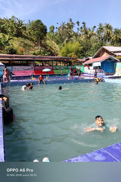The image depicts a group of people swimming and playing in a pool at an outdoor recreational facility. The pool is bordered with blue tiles. Various people are visible inside the pool, including a boy swimming towards the camera in the foreground. Other individuals are standing at the pool's edge and swimming in the background. Behind the pool, there's a covered structure with colorful painted murals on the side. The setting appears to be in a rural or semi-rural area, with trees and vegetation covering a hillside in the background. Based on the timestamp, the photo was taken on May 30, 2026 at 09:52. The city is identified as Palohseulimeng, Indonesia.