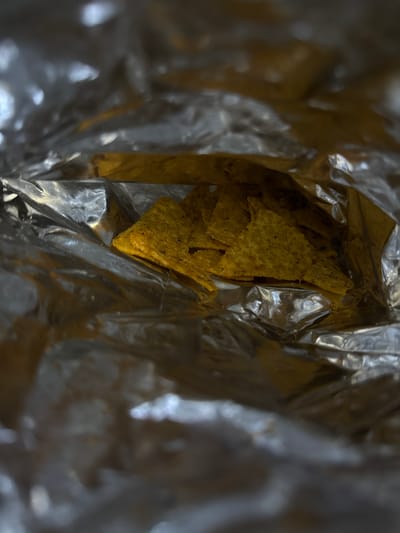 This close-up, overhead shot depicts a crumpled bag of tortilla chips, with several chips visible inside. The chips are triangular and appear to be flavored, possibly with spices. The bag is made of a shiny, metallic material, common for snack packaging, and it is wrinkled and creased, suggesting it has been opened. The lighting creates highlights and shadows on the foil, adding texture to the scene. The image is focused on the chips and the interior of the bag, creating an intimate perspective. There is no text visible in the image. The setting is not discernible beyond the immediate context of the snack bag. There are no people, specific time of day, weather conditions, or location cues to suggest it's in Getafe, Spain.