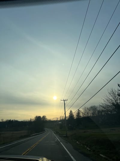 The image displays a rural road scene in the Town of New Windsor, United States of America, captured from the interior of a vehicle. A low, bright sun is positioned slightly left of the image center against a sky exhibiting a gradient from pale yellow near the horizon to light blue and grey overhead, with dispersed cloud cover. Six dark, parallel power lines extend diagonally across the upper right quadrant of the frame. A wooden utility pole stands on the right side of the asphalt road, supporting these lines. The two-lane road features a solid white line on the right shoulder and double yellow center lines, curving towards the horizon. To the left of the road, a metal guardrail runs parallel to dry, sparse vegetation, succeeded by a dense line of bare deciduous trees and some evergreens. A small, distant white structure is partially visible behind the trees on the left. On the right, a grassy verge with a ditch borders the road, leading to a rising embankment densely covered with bare deciduous and evergreen trees. A mailbox on a post is visible near the utility pole on the right. No individuals are depicted.
