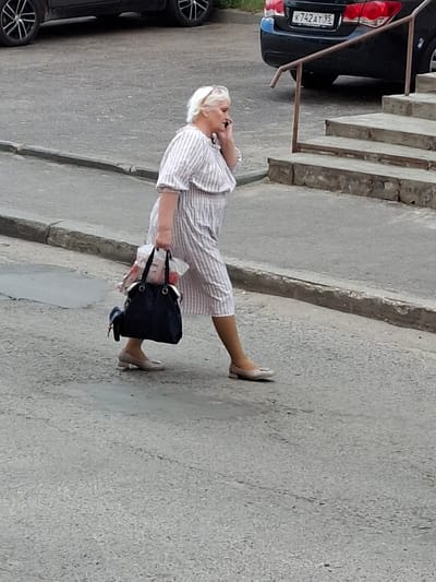 This is a vertical, full-body, eye-level shot of an older woman walking outdoors. She has blonde hair and is wearing a light-colored, pinstriped dress. She is holding a mobile phone to her ear with her right hand and is carrying a dark blue handbag over her left shoulder. Her left leg is extended forward, and her right leg is bent at the knee. She appears to be walking on a concrete sidewalk next to a set of outdoor stairs, with a parked black car on the left side of the frame. The car's license plate is partially visible, displaying the text "K742AT 95". The scene suggests it is daytime, with ample natural light. The overall setting appears to be an urban or suburban environment in Bel'kovo, Russia. There are no overt indications of the woman's emotional state.
