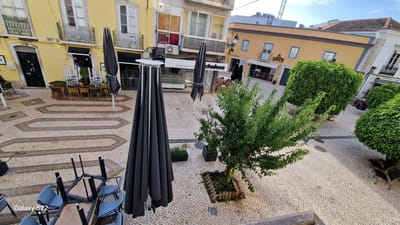 The image depicts an outdoor seating area in a European city square, likely in Faro, Portugal, given the architectural style and the "Galaxy S22" watermark. The square is paved with cobblestones and features decorative geometric patterns. Several restaurants or cafes are visible along the buildings that surround the square, with awnings and shopfronts.

In the foreground, a table with chairs is overturned, suggesting it might have been closed for the day or due to weather. Two large black umbrellas are also present, one partially obstructing the view, and the other in the mid-ground. Stacks of chairs and tables are neatly arranged against the buildings, indicating preparations for opening or closure.

A small tree is planted in a raised bed in the mid-ground, surrounded by a wooden border. Further back, several large, neatly trimmed evergreen shrubs add greenery to the square. In the distance, a few individuals are seated in the shade of the shrubs, their presence minimal and non-intrusive to the overall scene. The sky is partly cloudy, with a clear blue sky visible towards the top right. The overall impression is one of a quiet, European urban space, possibly during off-peak hours.