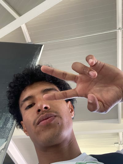 A close-up, tilted shot focuses on a young man with curly black hair and tan skin. He's positioned to the right, looking downward with a slight smirk. A hand, likely his own, is held above his face, making a "V" sign with the index and middle fingers. The background consists of a white ceiling with parallel lines and a partially visible black surface along the lower left edge. He is wearing a green-trimmed shirt.