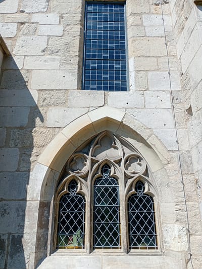 The image shows a close-up view of a stone building with two windows, located in Walkenried, Germany. The upper window is a tall, narrow rectangle with a grid of small, dark panes. The lower window is Gothic in style, featuring an arched top with ornate tracery and three smaller, diamond-paned windows within. The stonework of the building is rough and appears aged, with some shadows cast from an unseen object or structure to the left. A thin, light-colored wire runs vertically along the right side of the stone wall, attached at intervals by small fixtures. The overall impression is of architectural detail on an old building.