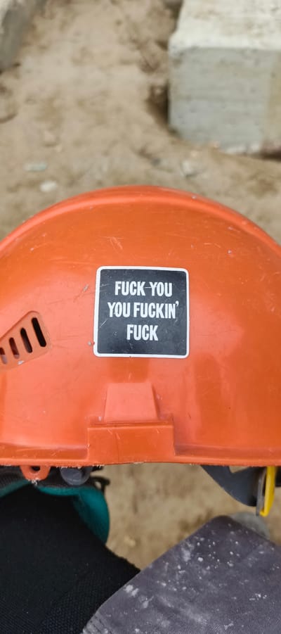 The image shows an orange hard hat with a sticker on it that says "FUCK YOU YOU FUCKIN' FUCK". The hard hat is on the head of a person wearing dark clothing, likely work clothes, with some white dust or debris on them. The background appears to be a construction site with sand and concrete blocks. There is no indication of the specific location within Primorye, Russia.