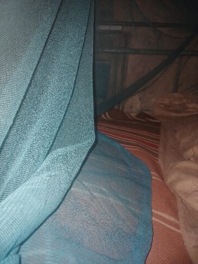 A close-up shot focuses on teal netting, its texture visible. In the background, indistinct bedding with stripes and soft textures adds depth. A dark structure, seemingly a frame or support, is subtly present behind the netting. The lighting emphasizes the fabric's weave and creates contrasting shadows. The composition features varying textures and a muted color palette, emphasizing fabric details.
