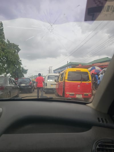 The image, taken from inside a vehicle, shows a traffic scene in what appears to be Benin City, Nigeria. The cracked windshield obscures part of the sky. A red and yellow minibus with the license plate "ANG-21XB" is directly in front. Several other vehicles are visible, including a white van and black cars. A man wearing a red shirt is walking between the vehicles. In the background, there are commercial buildings and an umbrella. Power lines stretch across the sky.