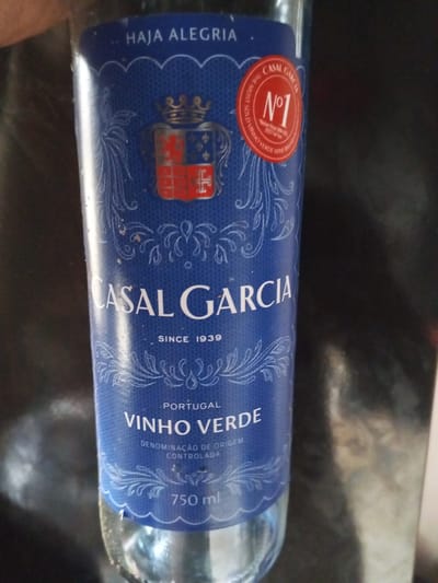 A close-up shot shows a blue label on a glass bottle of wine. The label features a crest with a crown, a red and blue shield, and decorative scrollwork. Text on the label reads "HAJA ALEGRIA," "CASAL GARCIA," "SINCE 1939," "PORTUGAL," "VINHO VERDE," and "DENOMINAÇÃO DE ORIGEM CONTROLADA." A circular red sticker on the upper right of the label states "CASAL GARCIA N°1 THE MOST SOLD VINHO VERDE WINE." The bottle is partially filled with a clear liquid and is standing upright against a dark, reflective background. The environment appears to be indoors.