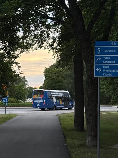 The image shows a blue bus driving on a wet asphalt road, likely after rain, with a tree in the foreground and a grassy area on the right. The sky in the background is overcast with streaks of yellow and white light, suggesting it might be dusk or dawn.
The bus has text on its side, including "Kofimatkallasi mikana" and "PORIN YH-ASUNNOT". There is also a road sign visible with Finnish place names like "Tuorsniemi", "Yyteri", "Karjaranta", "Luvia", "Urheilukesku", and "Käppärä", indicating the location is Pori, Finland. A blue roundabout sign is also visible near the road. The overall scene appears to be a quiet, perhaps rural, road with natural elements like trees and grass dominating the landscape. The lighting suggests a subdued atmosphere. There are no people clearly visible in the image.